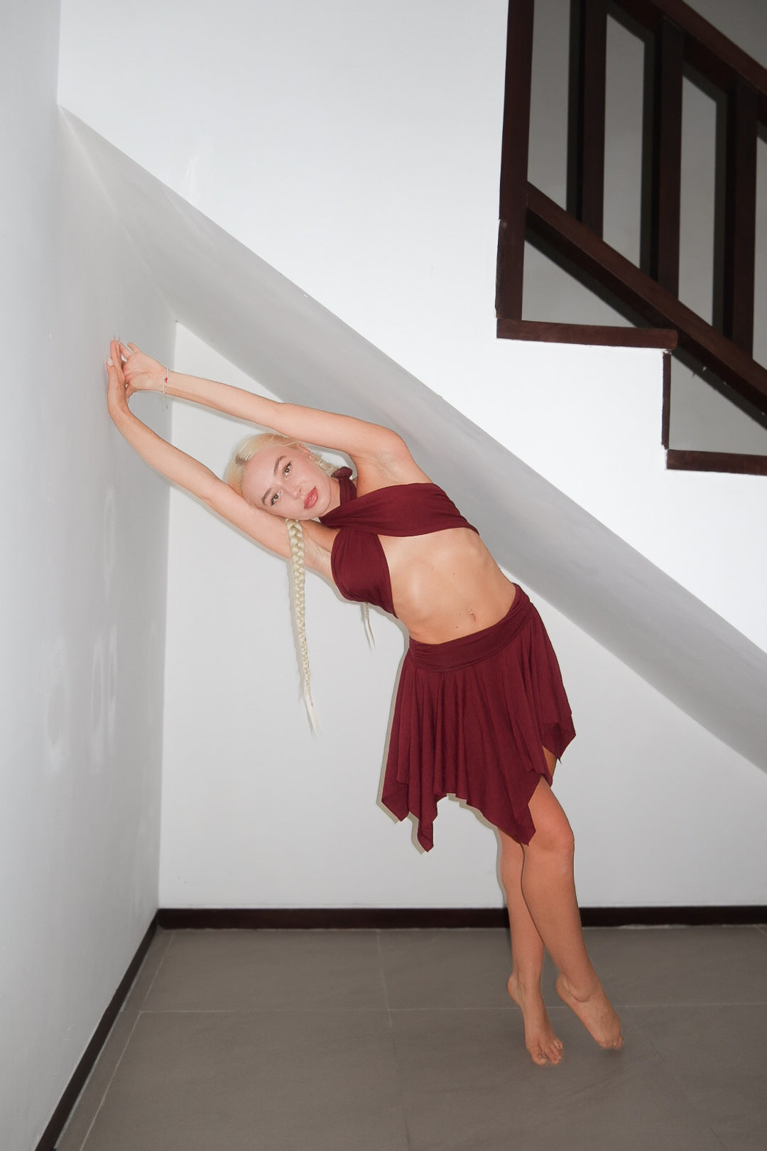 Woman in a burgundy dress posing in a room with white walls and a staircase.